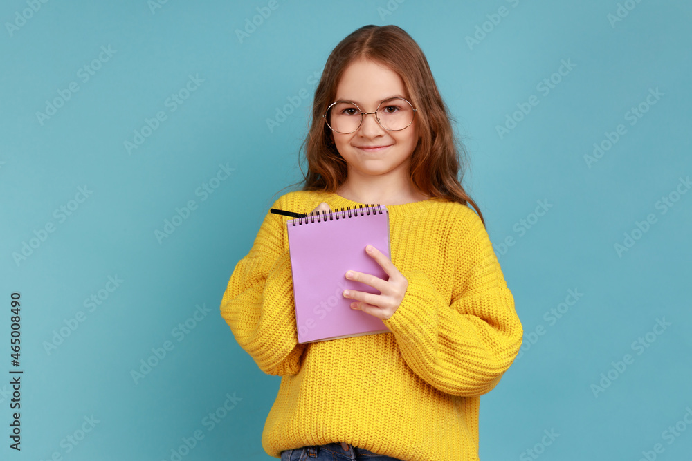 Portrait of positive little girl writing in notebook, looks smiling at camera, child doing homework, wearing yellow casual style sweater. Indoor studio shot isolated on blue background. Portrait of positive little girl writing in notebook, looks smiling at camera, child doing homework, wearing yellow casual style sweater. Indoor studio shot isolated on blue background.