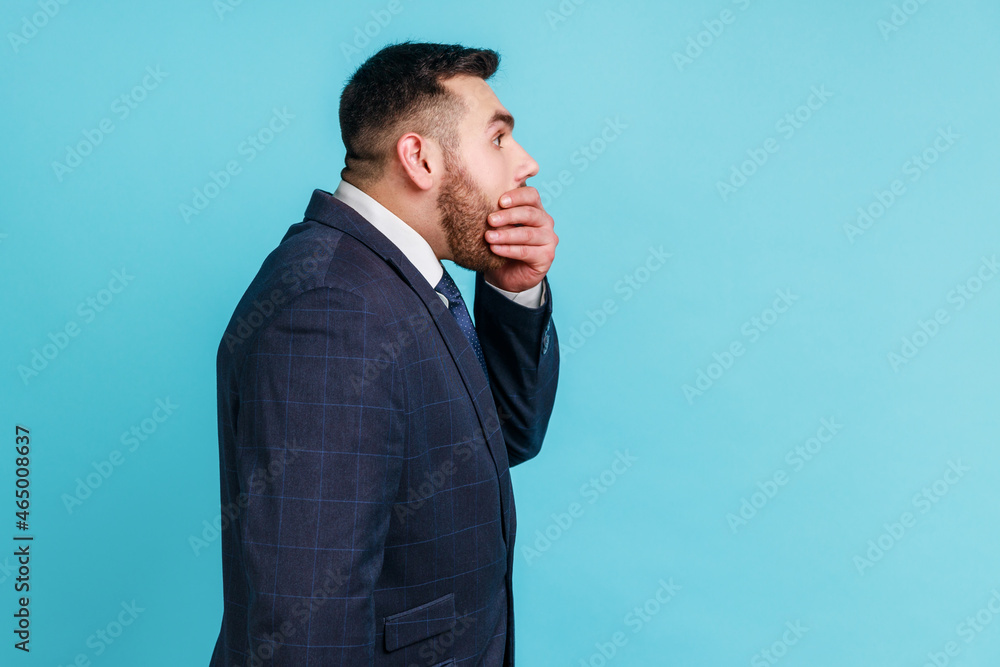 I won't say anyone! Side view of bearded man wearing official style suit covering mouth with hand, keeping terrible secret truth, don't want to talk. Indoor studio shot isolated on blue background. I won't say anyone! Side view of bearded man wearing official style suit covering mouth with hand, keeping terrible secret truth, don't want to talk. Indoor studio shot isolated on blue background.