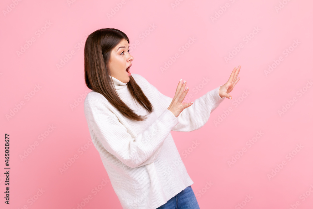 Side view portrait of scared female with raised arms, sees something awful, afraids, stop gesture, wearing white casual style sweater. Indoor studio shot isolated on pink background. Side view portrait of scared female with raised arms, sees something awful, afraids, stop gesture, wearing white casual style sweater. Indoor studio shot isolated on pink background.