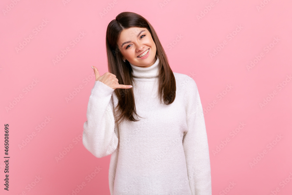 Smiling woman standing with happy playful expression, making telephone gesture with hand flirting, wearing white casual style sweater. Indoor studio shot isolated on pink background. Smiling woman standing with happy playful expression, making telephone gesture with hand flirting, wearing white casual style sweater. Indoor studio shot isolated on pink background.