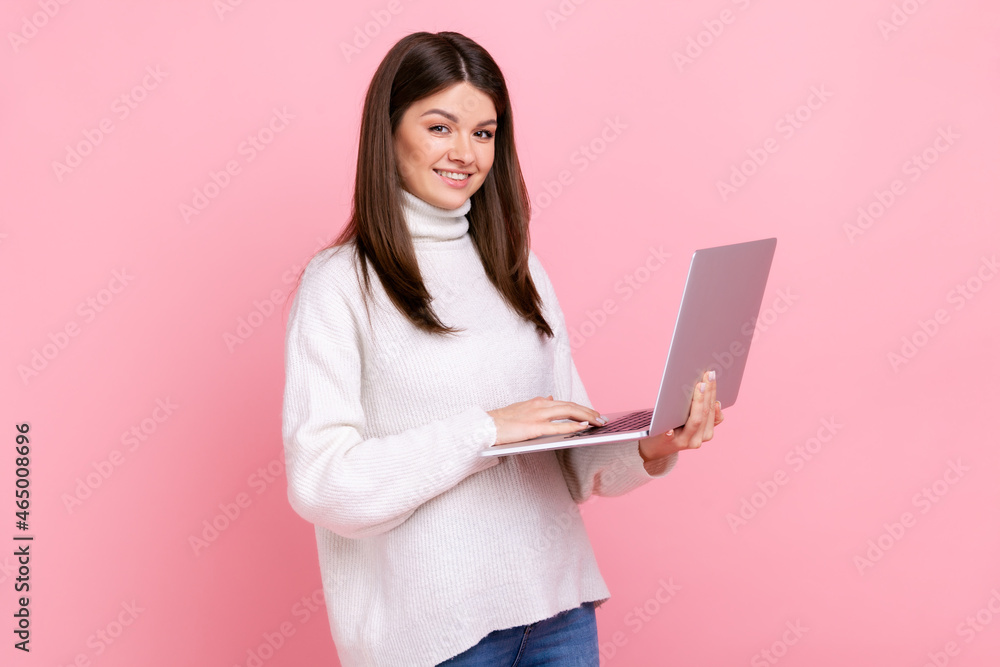 Successful office worker girl looking at camera with happy face, holding laptop, typing on keyboard, wearing white casual style sweater. Indoor studio shot isolated on pink background. Successful office worker girl looking at camera with happy face, holding laptop, typing on keyboard, wearing white casual style sweater. Indoor studio shot isolated on pink background.