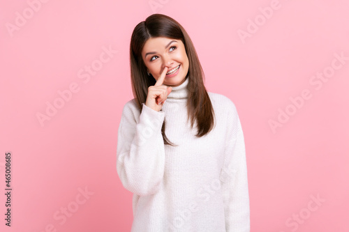 Female touching nose, showing liar gesture, angry about falsehood, outright deception, fake news, wearing white casual style sweater. Indoor studio shot isolated on pink background.