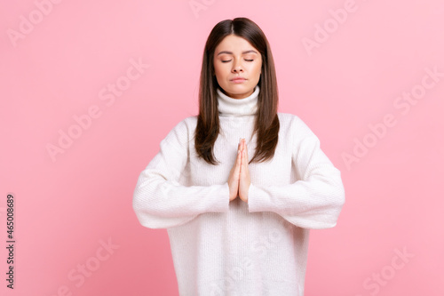 Calm relaxed female standing with closed eyes, breathing, doing yoga exercising, feeling harmony, wearing white casual style sweater. Indoor studio shot isolated on pink background.