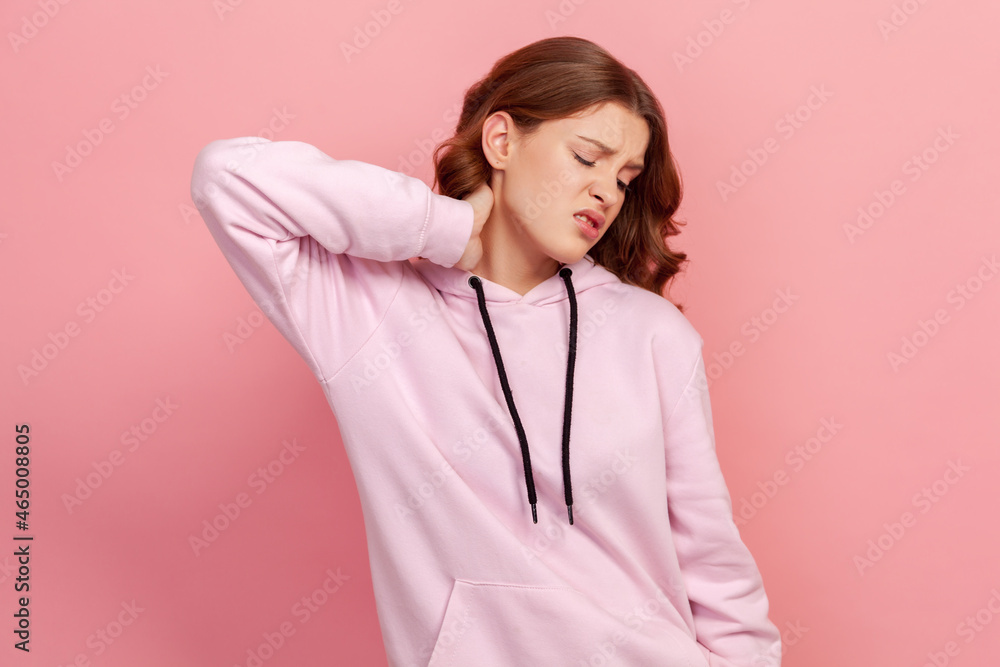 Portrait of upset brunette teen girl in hoodie touching neck feeling pain and numbness, worried about muscle tension, osteochondrosis. Indoor studio shot isolated on pink background Portrait of upset brunette teen girl in hoodie touching neck feeling pain and numbness, worried about muscle tension, osteochondrosis. Indoor studio shot isolated on pink background