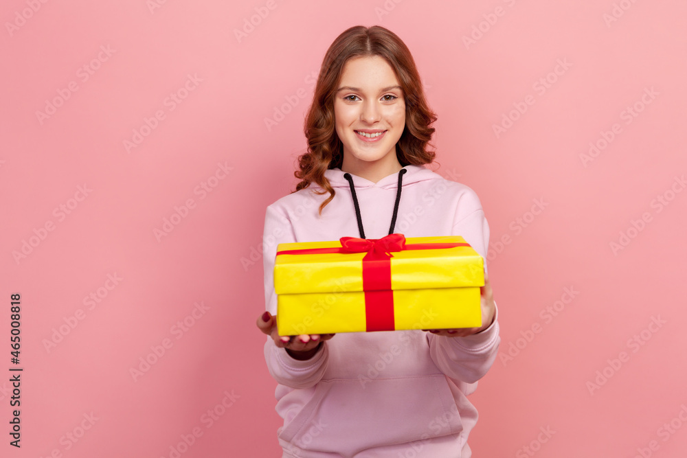 Portrait of happy curly haired teen girl in hoodie presenting wrapped gift box and looking at camera with smile, holiday greeting. Indoor studio shot isolated on pink background Portrait of happy curly haired teen girl in hoodie presenting wrapped gift box and looking at camera with smile, holiday greeting. Indoor studio shot isolated on pink background