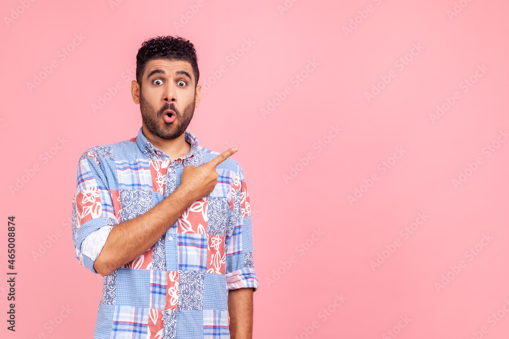 Excited surprised handsome man with beard wearing blue casual shirt looking at camera with open mouth, pointing finger aside, showing copy space. Indoor studio shot isolated on pink background. Excited surprised handsome man with beard wearing blue casual shirt looking at camera with open mouth, pointing finger aside, showing copy space. Indoor studio shot isolated on pink background.