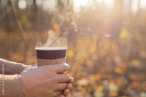 A man holds a disposable paper cup with a drink in the park in the fall. The concept of drinks, tea, coffee, autumn, warm up, rest.