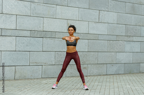 Young flexible woman in top and leggings does sports exercises for arms training near light grey stone wall