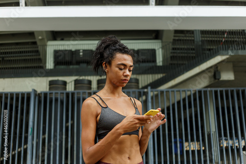 Young sportswoman in stylish top looks at modern smartphone resting during training on empty road against building metal gate