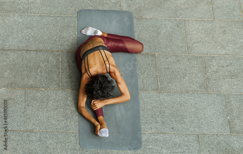 Slim biracial lady in stylish tracksuit does stretching exercises for legs on mat at training on city pavement view from above