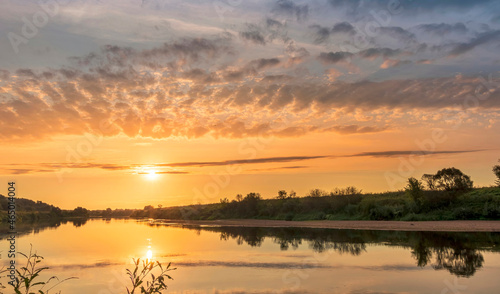 Scenic view at beautiful summer river sunset with reflection on water with green bushes  calm water  deep colorful cloudy sky and glow on horizon on a background  spring evening landscape