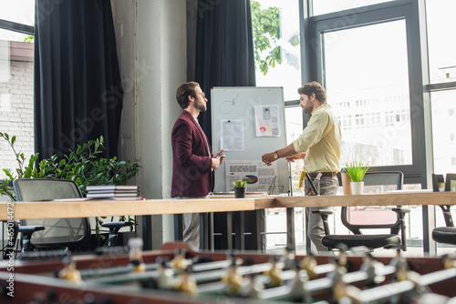 Side view of businessmen working with papers on flipchart near blurred table football in office