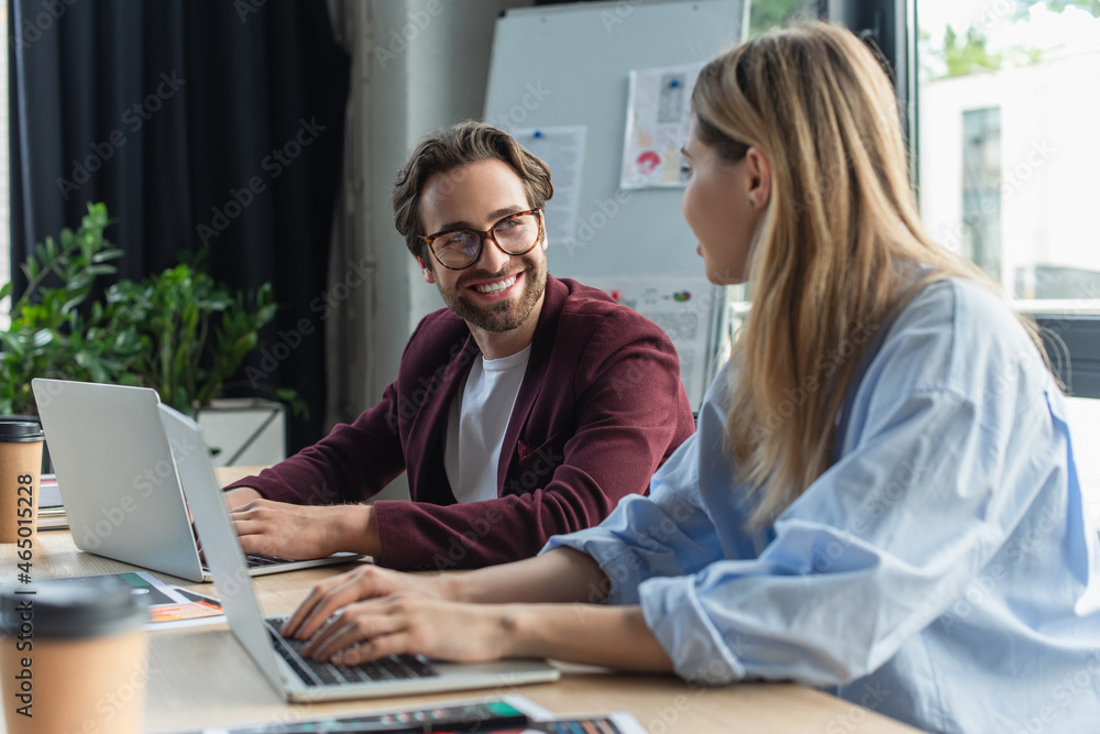 Smiling businessman looking at blurred colleague using laptop in office Smiling businessman looking at blurred colleague using laptop in office