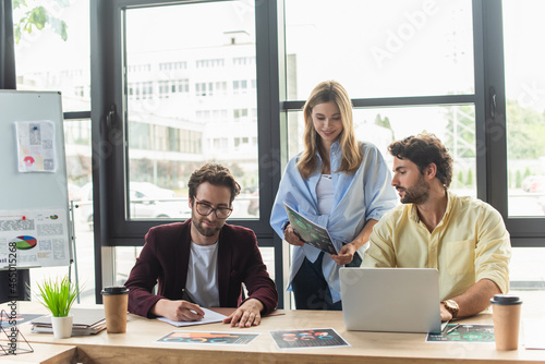 Smiling businesswoman holding document near businessmen with laptop in office