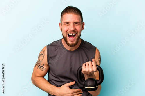 Young caucasian man holding ketlebell isolated on blue background laughing and having fun. photo