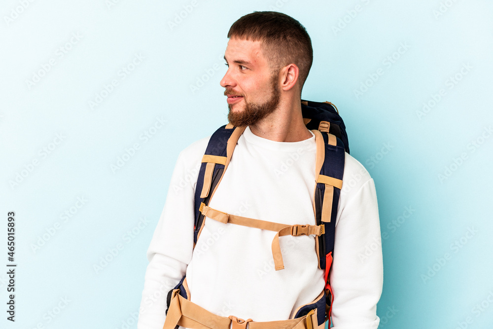 Young hiker caucasian man isolated on blue background looks aside smiling, cheerful and pleasant. Young hiker caucasian man isolated on blue background looks aside smiling, cheerful and pleasant.