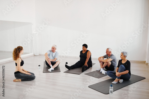 Senior people sitting on the floor and practicing yoga together with coach during yoga class in studio