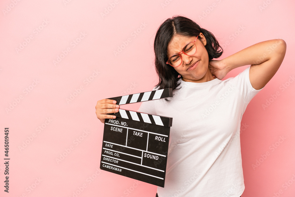Young latin woman holding clapperboard isolated on pink background touching back of head, thinking and making a choice.