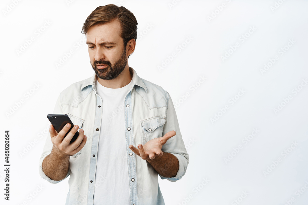 Guy looking confused at his smartphone and frowning puzzled, cant understand app, standing over white background Guy looking confused at his smartphone and frowning puzzled, cant understand app, standing over white background