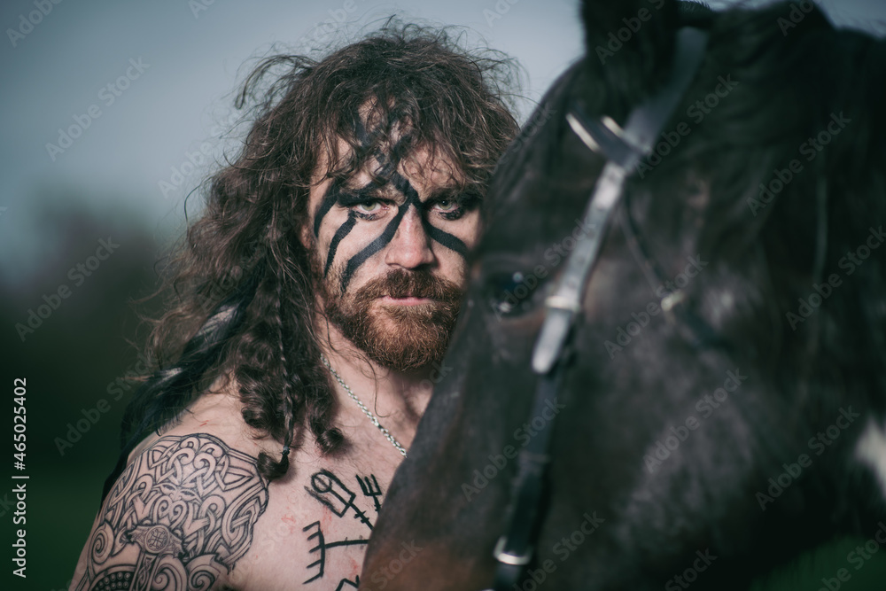 Obraz premium Medieval warrior berserk Viking portrait with tattoo on skin, cuts on body and braids in hair with weapons to attacks enemy. Concept historical photo