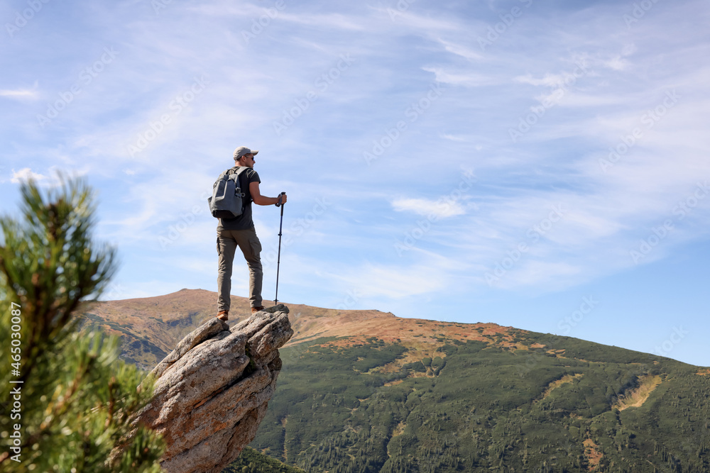 Man with backpack and trekking poles on rocky peak in mountains, back view