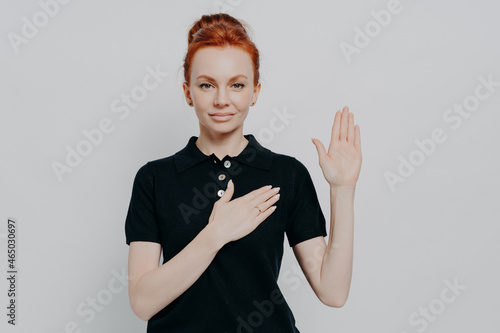 tudio shot of serious red haired female making promise, swearing with hand on chest in studio photo