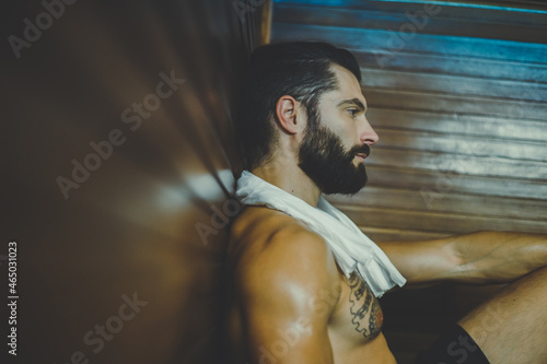 Beautiful healthy boy relaxing in a woden sauna. Young bearded man resting after workout in the gym. Fitness male with tatoo and white towel, laying on the bench in the spa. Wellness, health concept