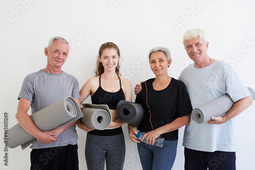 Portrait of young trainer smiling at camera while standing with senior people with exercise mats against the white wall