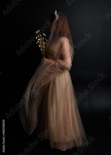 Full length portrait of red haired girl wearing a creamy fantasy gown like a fairy goddess costume. standing pose with elegant gestural movement backwards the the camera, isolated on dark studio 