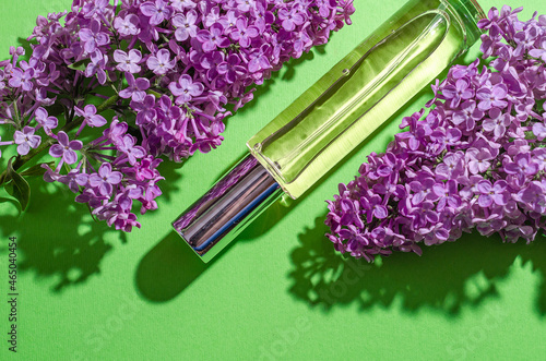 Purple lilac flowers and a glass bottle with perfume on a green background. Bright shadows and light. Place for your text. Flat lay.