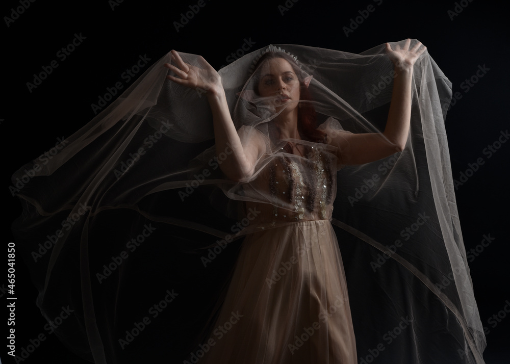 Naklejka premium Full length portrait of red haired girl wearing a creamy fantasy gown like a fairy goddess costume. standing pose with elegant gestural movement , isolated on dark studio background.