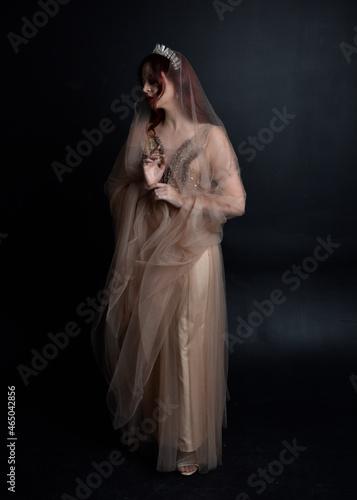 Full length portrait of red haired girl wearing a creamy fantasy gown like a fairy goddess costume. standing pose with elegant gestural movement , isolated on dark studio background.