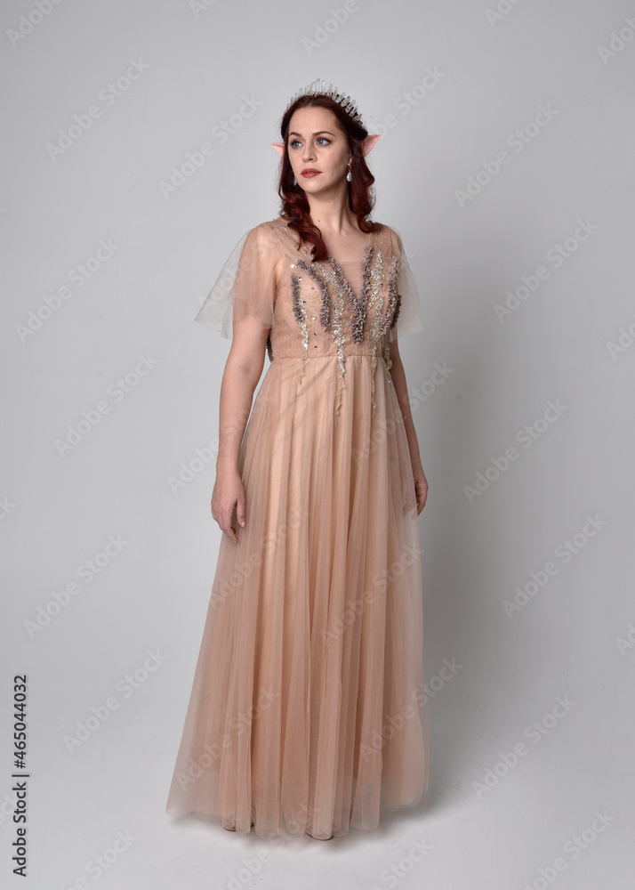 Full length portrait of red haired girl wearing a creamy fantasy gown and crystal crown, like a fairy goddess costume. standing pose with elegant gestural arm movement , isolated on light studio b Full length portrait of red haired girl wearing a creamy fantasy gown and crystal crown, like a fairy goddess costume. standing pose with elegant gestural arm movement , isolated on light studio b
