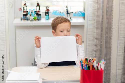A cute boy first grader in a school uniform at home while isolated at his desk holds a sheet of blank paper. Selective focus. Portrait