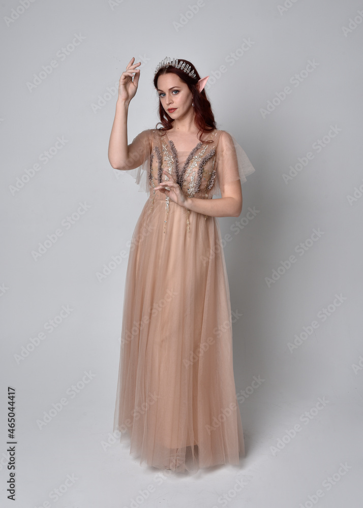 Full length portrait of red haired girl wearing a creamy fantasy gown and crystal crown, like a fairy goddess costume. standing pose with elegant gestural arm movement , isolated on light studio b Full length portrait of red haired girl wearing a creamy fantasy gown and crystal crown, like a fairy goddess costume. standing pose with elegant gestural arm movement , isolated on light studio b