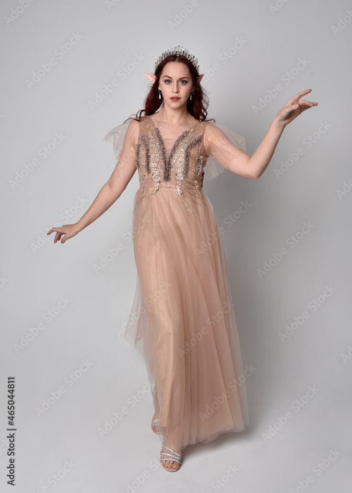 Full length portrait of red haired girl wearing a creamy fantasy gown and crystal crown, like a fairy goddess costume. standing pose with elegant gestural arm movement , isolated on light studio b Full length portrait of red haired girl wearing a creamy fantasy gown and crystal crown, like a fairy goddess costume. standing pose with elegant gestural arm movement , isolated on light studio b