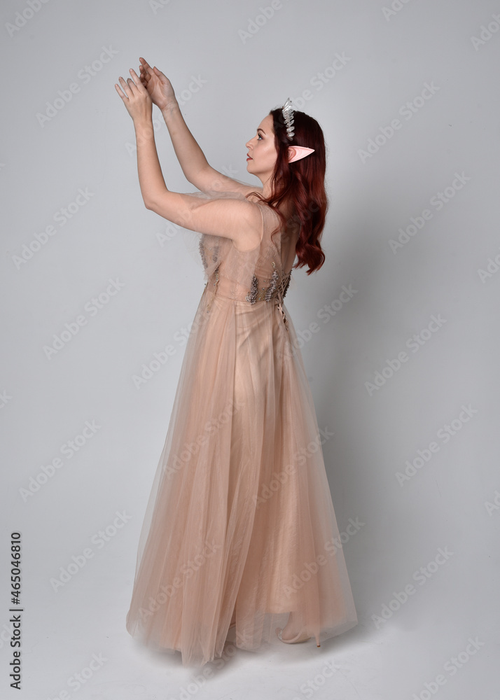 Full length  portrait of red haired  girl wearing a creamy fantasy gown and crystal crown, like a fairy goddess costume.  standing  pose with elegant gestural arm movement , isolated on light studio b