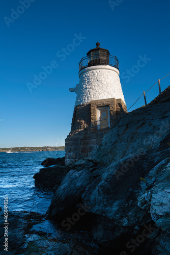 White lighthouse on the rocky cliff over the blue ocean water against clear sky. Castle Hill Lighthouse in Newport, Rhode Island. © Naya Na