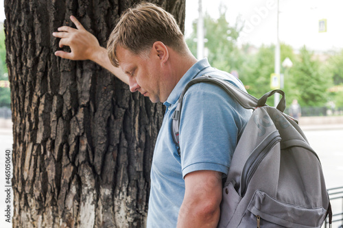Photos adult man with a backpack leaned his hand on a tree feeling ill on the street