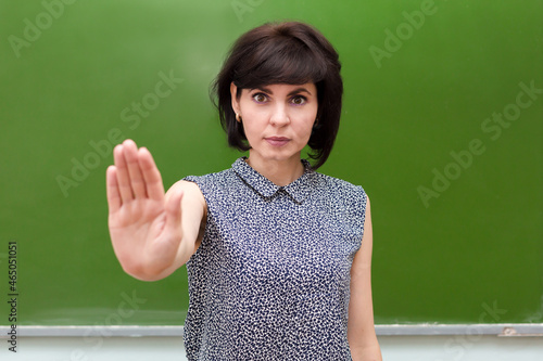 A strict teacher at the blackboard in a school classroom shows a stop sign to violence at school with her hand.