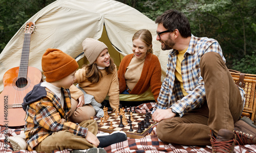 Happy smiling family playing chess game at campsite during camping trip in nature