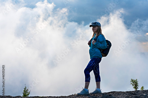 Young woman in cap and blue sporty clothes with backpack standing on the edge of mountain against background of white clouds, hiking and active healthy lifestyle concept tourism