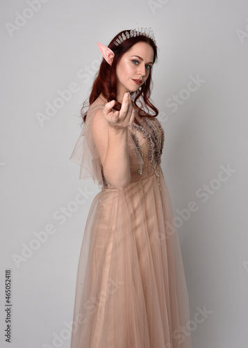 Full length portrait of red haired girl wearing a creamy fantasy gown and crystal crown, like a fairy goddess costume. standing pose with elegant gestural hands, isolated on studio background