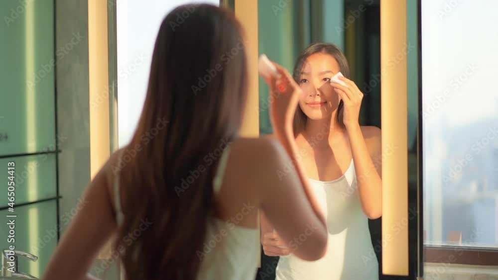 Beautiful young Asian woman looking in mirror of hotel bathroom and cleaning face with morning sunlight. Femininity concept Beautiful young Asian woman looking in mirror of hotel bathroom and cleaning face with morning sunlight. Femininity concept