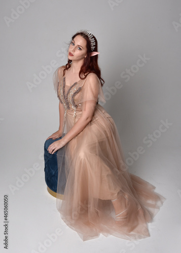 Full length portrait of red haired girl wearing a creamy fantasy gown and crystal crown, like a fairy goddess costume. sitting pose with elegant gestural hands, isolated on light studio background.