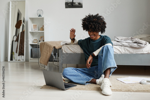Portrait of teenage African-American boy sitting on floor at home or in college dorm and waving to camera wile speaking by video chat online, copy space