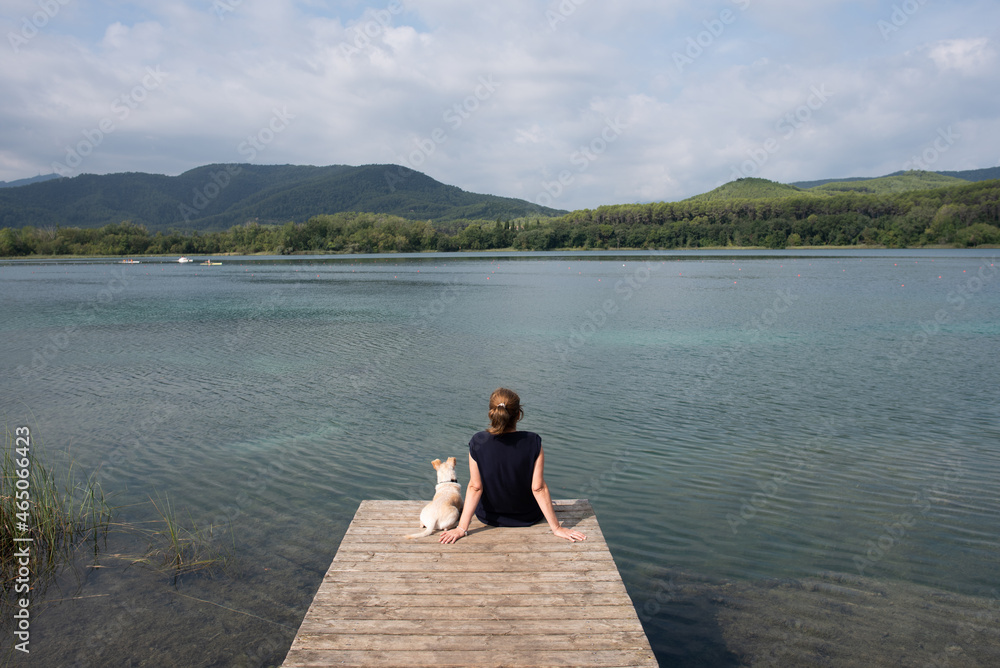 Adult woman and her pet sitting on the wooden dock looking towards the Banyoles lake, in Girona