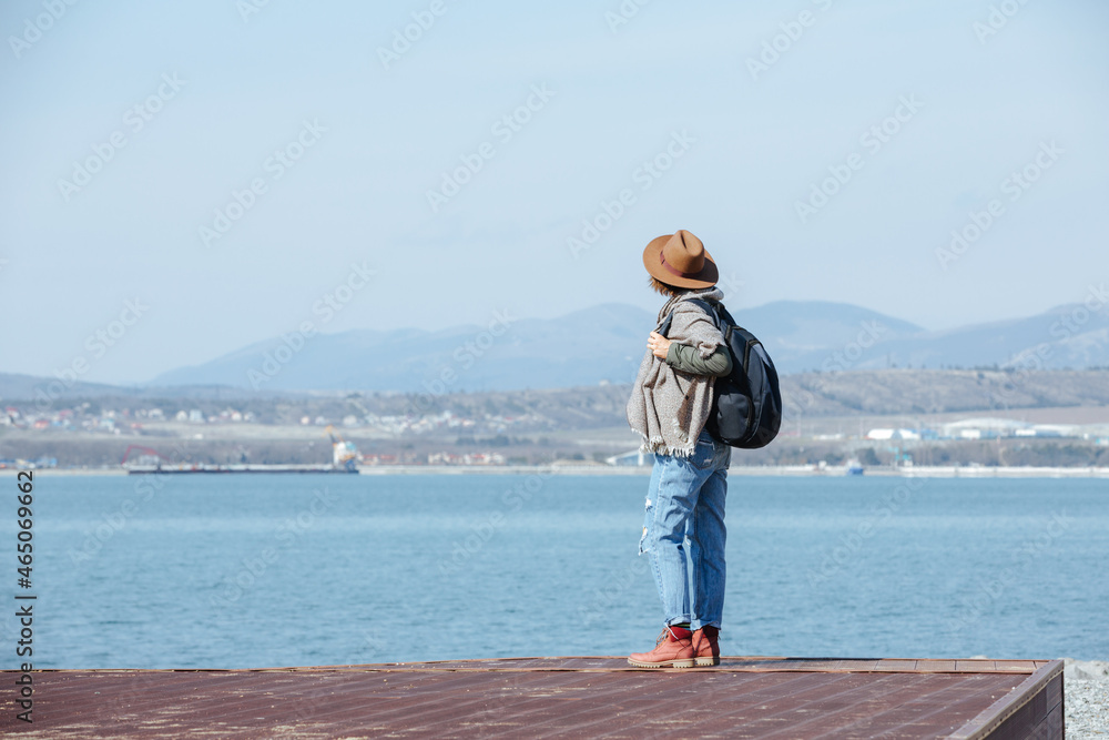 Stylish modern woman traveler with a backpack. Stands and enjoys the view of the mountains and the sea. Cool weather and sunny day.