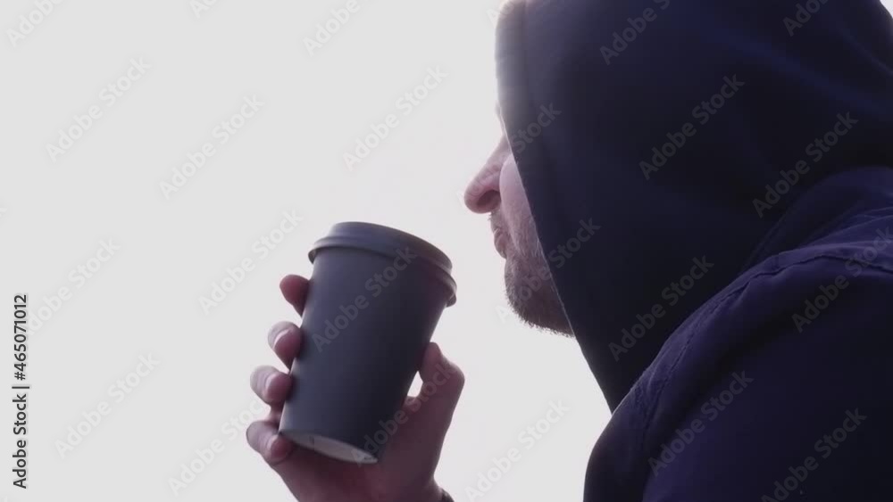 A man of European appearance in a dark blue sweatshirt with a hood is drinking coffee in the park on a bench. Close-up of a man drinking coffee outdoors A man of European appearance in a dark blue sweatshirt with a hood is drinking coffee in the park on a bench. Close-up of a man drinking coffee outdoors