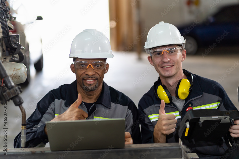 Smiling engineer worker working with computer notebook for control automatic robotic arm machine in the factory. Male technician worker maintenance automatic robotic hand in smart factory Smiling engineer worker working with computer notebook for control automatic robotic arm machine in the factory. Male technician worker maintenance automatic robotic hand in smart factory